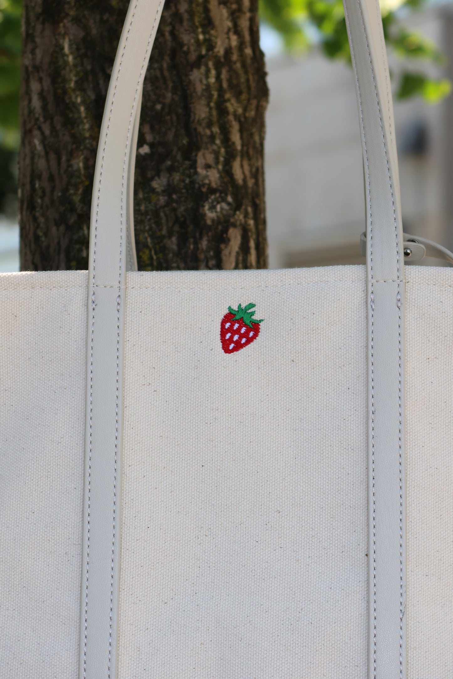 White tote bag with a red and green embroidered strawberry design on a tree branch.