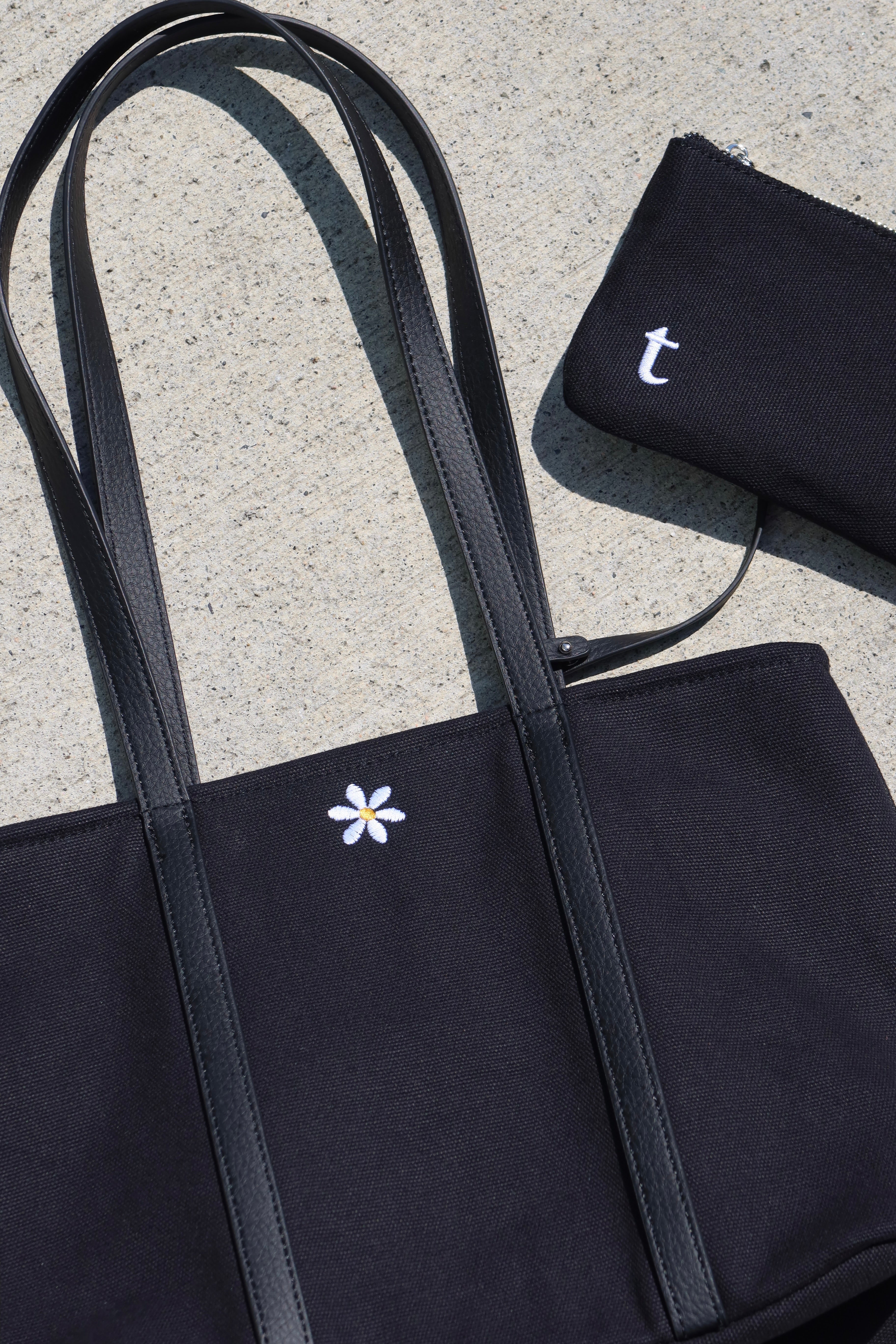 Black tote bag with a white flower design and matching pouch on a concrete surface. includes a small pouch that has interior credit card holder, and interior pockets. small flower embroidery, canvas tote bag