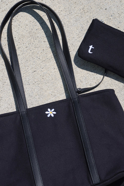 Black tote bag with a white flower design and matching pouch on a concrete surface. includes a small pouch that has interior credit card holder, and interior pockets. small flower embroidery, canvas tote bag