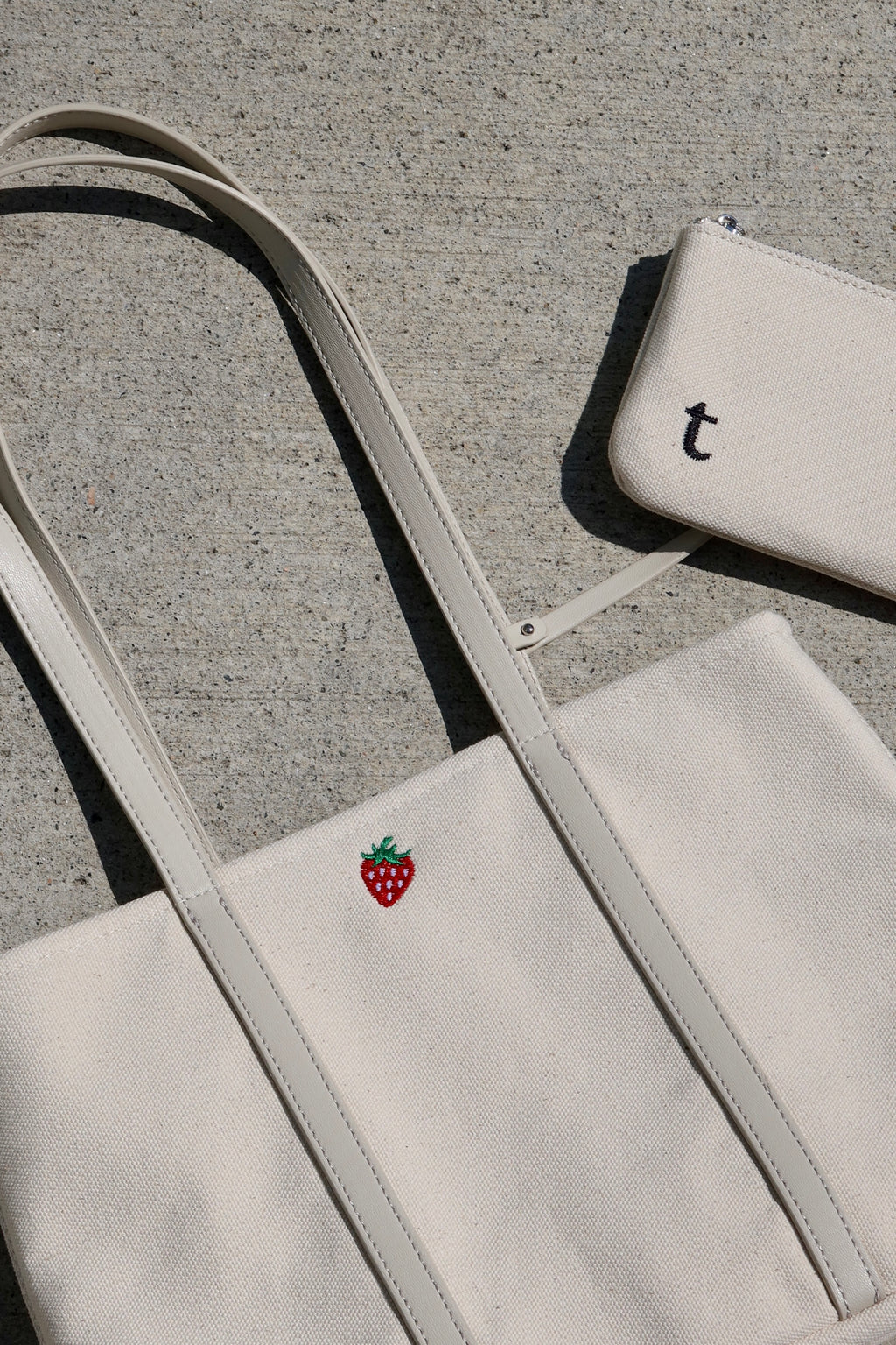 White tote bag with a red strawberry logo and matching pouch with built in credit card slots. placed on a concrete ground