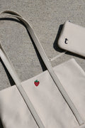 White tote bag with a red strawberry logo and matching pouch with built in credit card slots. placed on a concrete ground