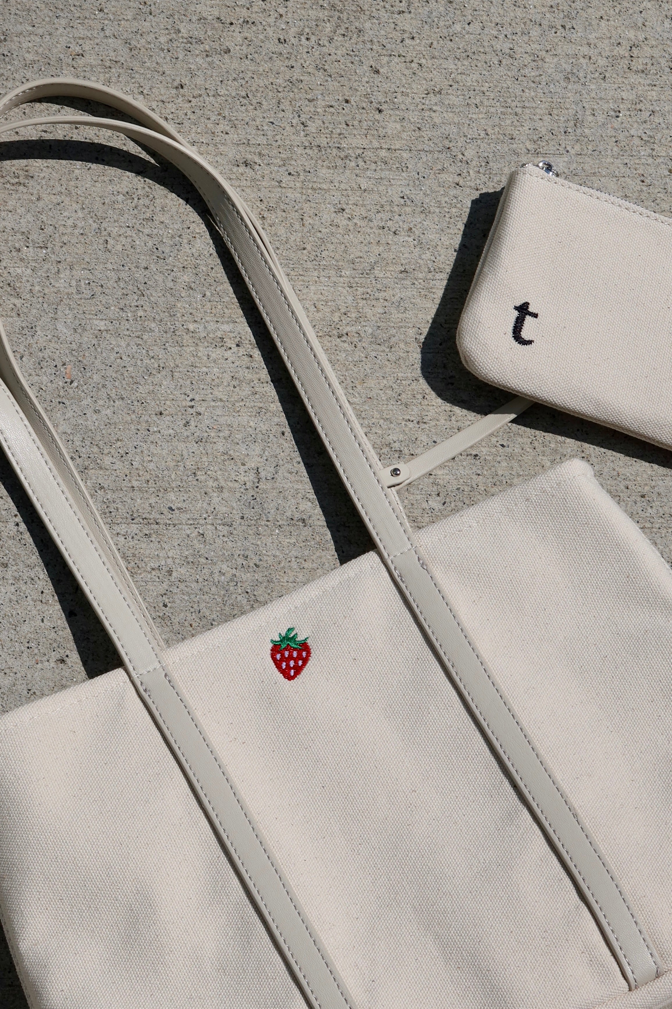 White tote bag with a red strawberry logo and matching pouch with built in credit card slots. placed on a concrete ground